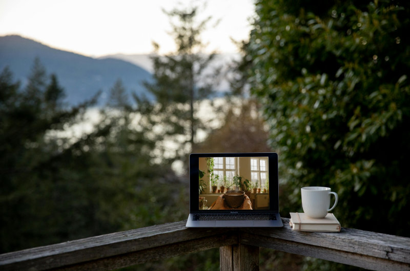 Laptop auf einem Holzgeländer inmitten der Natur, zeigt einen Kakao-Ritualraum auf dem Bildschirm – daneben eine Tasse und Bücher. Symbolbild für die Online-Begleitung mit Tanja Richter.