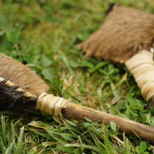 Eine handgefertigte schamanische Doppelrassel aus Rohhaut mit Holzgriffen und Lederschnur, liegend im grünen Gras