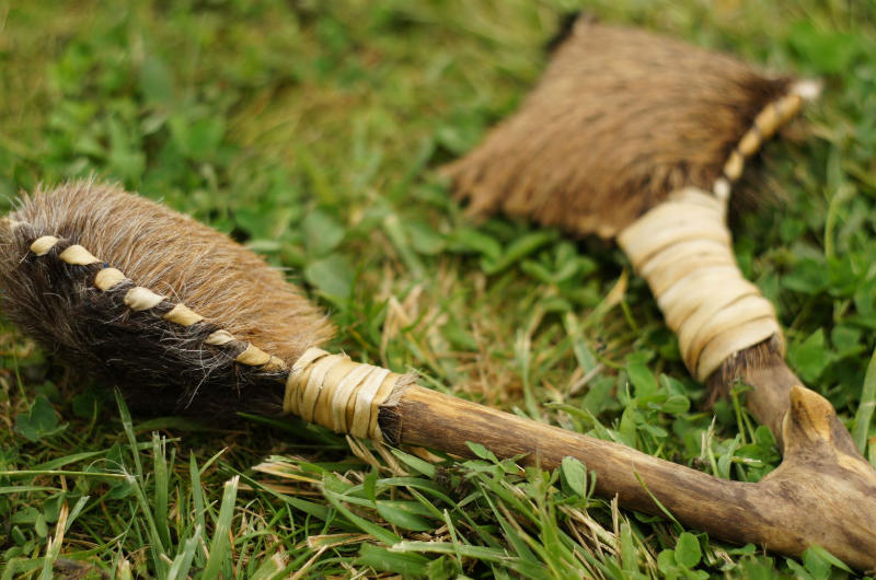 Eine handgefertigte schamanische Doppelrassel aus Rohhaut mit Holzgriffen und Lederschnur, liegend im grünen Gras