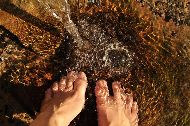 Zwei Füße im klaren, flachen Wasser auf steinigem Grund – symbolisch für Erdung, Reinigung und Verbindung mit Mutter Erde